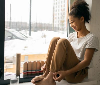 Full body side view of cheerful barefoot African American teenage girl in casual wear sitting near window in light room
