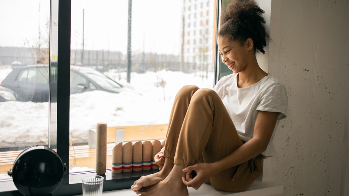 Full body side view of cheerful barefoot African American teenage girl in casual wear sitting near window in light room